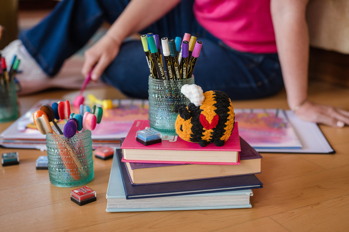 A cozy indoor scene with a small crocheted bee toy resting on a stack of books on a wooden floor. A person in a pink top and blue jeans is seated in the background, holding a pencil over art supplies, including colored pencils in textured glass cups and an artbook.
