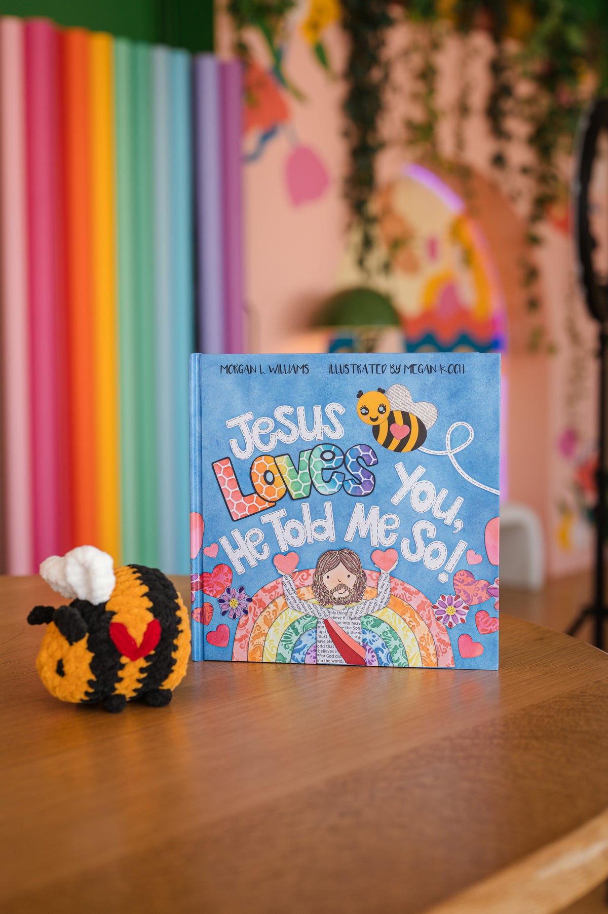 Children's book titled 'Jesus Loves You, He Told Me So!' with a bee plush toy on a wooden surface.