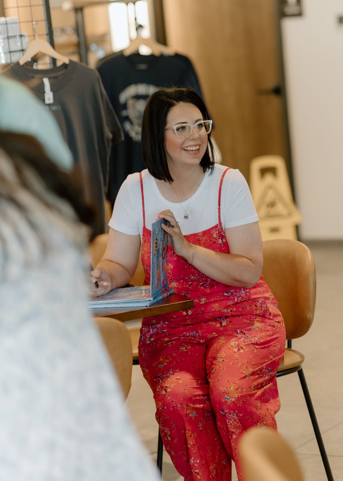 A woman with short dark hair and glasses sits in a cafe or boutique setting, smiling while holding a book or magazine. She is wearing a white t-shirt and a red floral overall jumpsuit.