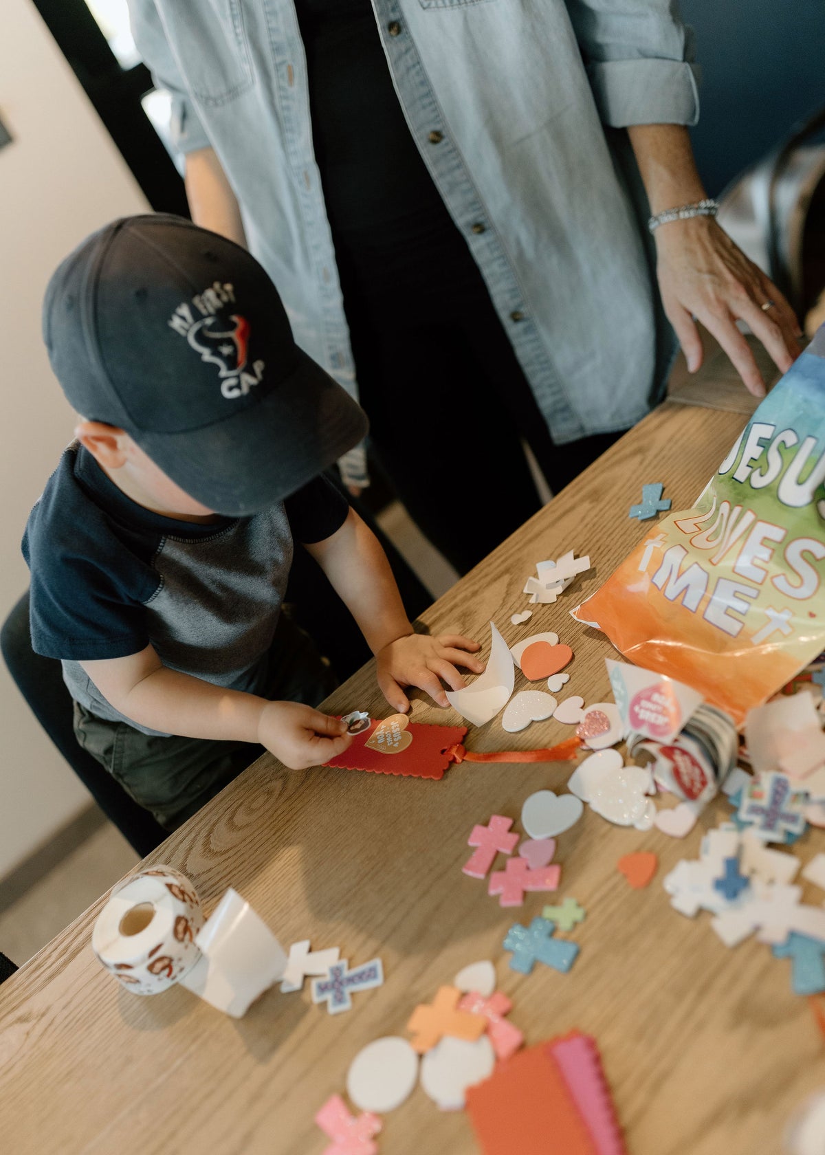 A young boy wearing a black baseball cap and a dark t-shirt sits at a wooden table, playing with colorful heart-shaped craft cutouts and a roll of patterned tape. An adult woman in a light denim shirt stands behind him.